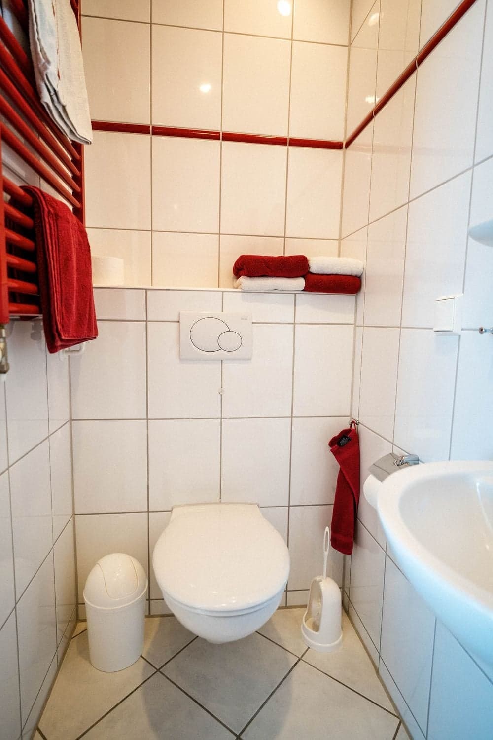 Weiß gefliestes Gästebad mit rotem Heizkörper, roten Handtüchern und Wand-WC