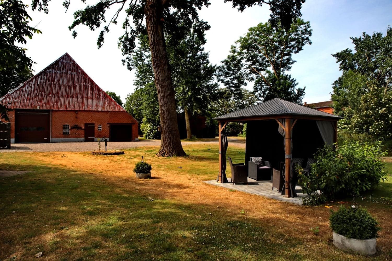 Rasenfläche mit Pavillon, großem Baum und der roten Scheune im Hintergrund