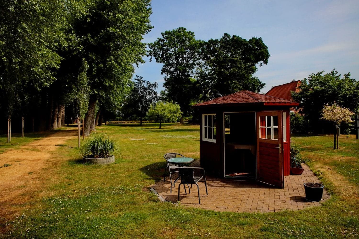 Rotes Gartenhäuschen mit geöffneter Tür, davor ein Tisch mit zwei Stühlen auf Pflaster